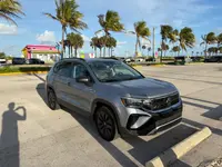 Silver 2023 Volkswagen Taos SUV, front passenger-side three-quarter view, parked in a beachside lot lined with palm trees