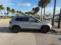 Silver 2023 Volkswagen Taos SUV in passenger-side profile, parked at a palm-lined marina with docked boats behind it