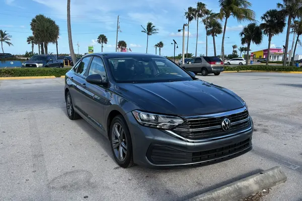 Gray 2024 Volkswagen Jetta sedan, front passenger-side three-quarter view, parked in a sunlit beachside lot lined with palm trees